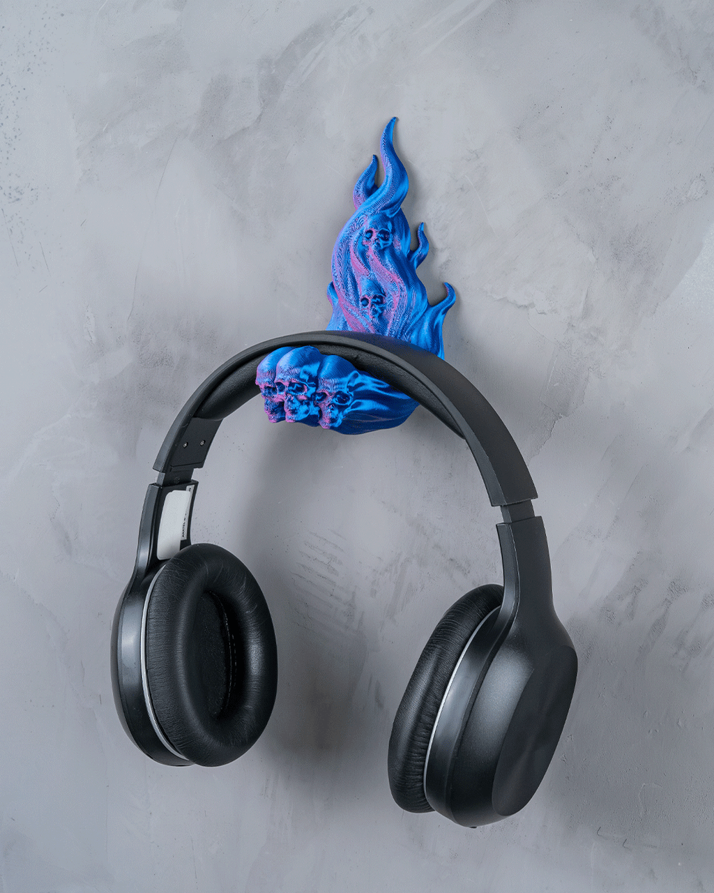 Skullflame Wand-Headset-Halter in Blau mit aufgehängtem Over-Ear-Kopfhörer, Frontalansicht an heller marmorierter Wand, Totenkopf-Reliefs im Flammen-Sockel sichtbar.