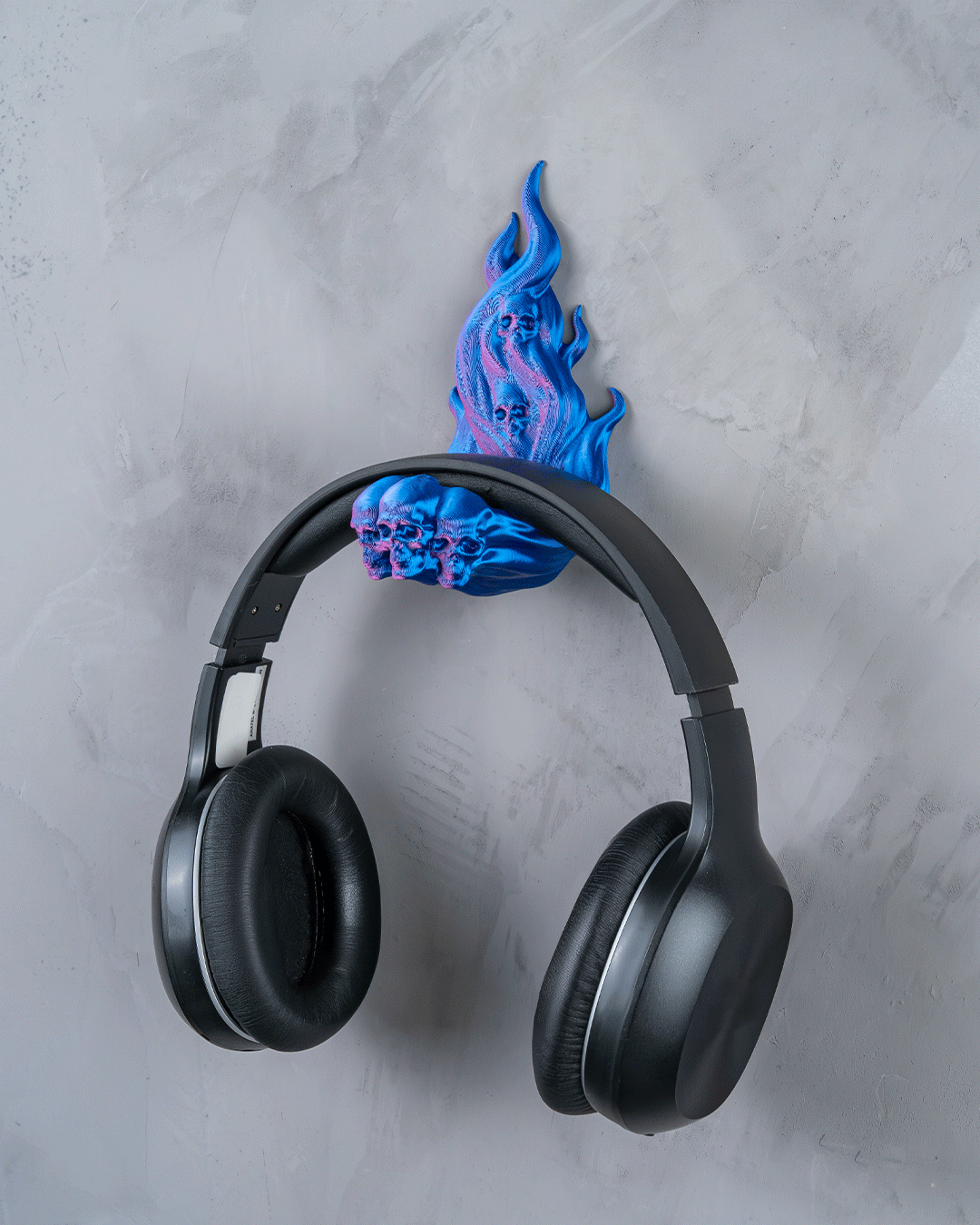 Skullflame Wand-Headset-Halter in Blau mit aufgehängtem Over-Ear-Kopfhörer, Frontalansicht an heller marmorierter Wand, Totenkopf-Reliefs im Flammen-Sockel sichtbar.