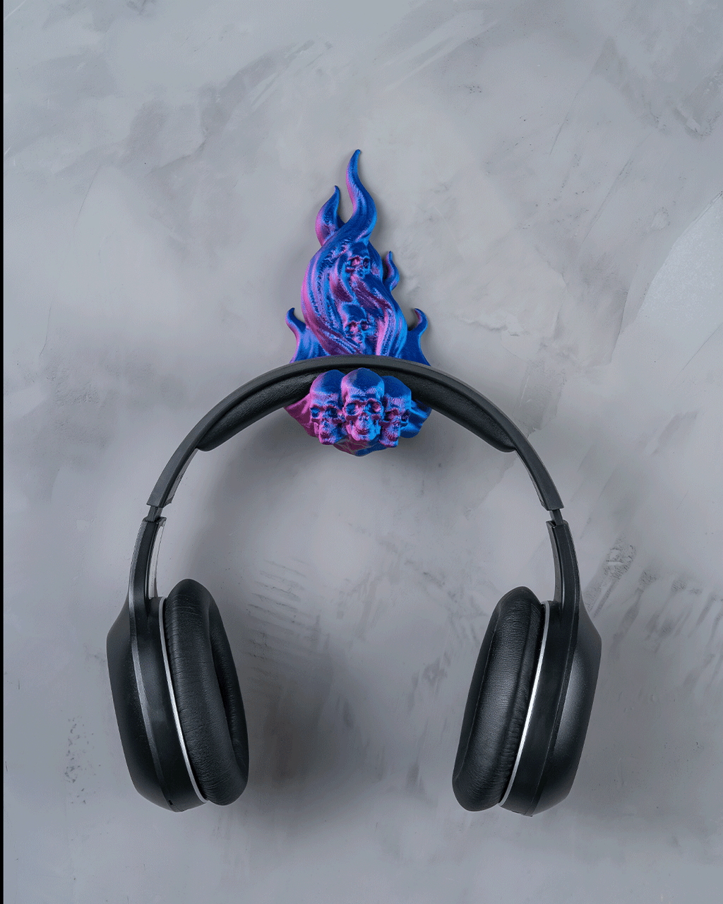 Skullflame Wand-Headset-Halter in Blau-Pink mit aufgehängtem Over-Ear-Kopfhörer, Frontalansicht an heller marmorierter Wand, Totenkopf-Reliefs im Flammen-Sockel sichtbar.