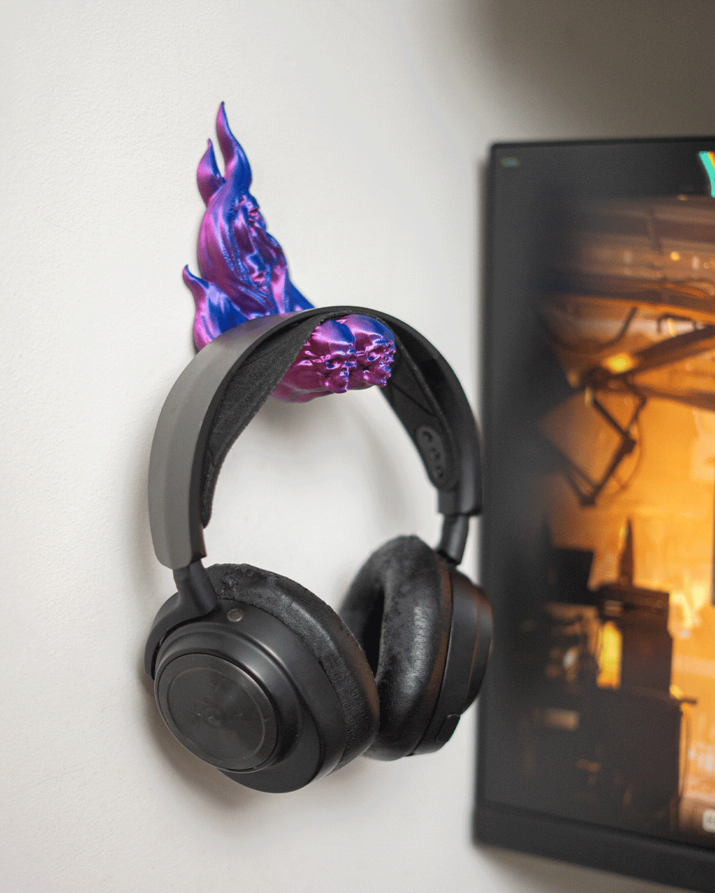 Skullflame Wand-Headset-Halter in Pink-Lila mit aufgehängtem Over-Ear-Kopfhörer, an heller Wand neben einem Monitor mit Spielszene im Hintergrund.