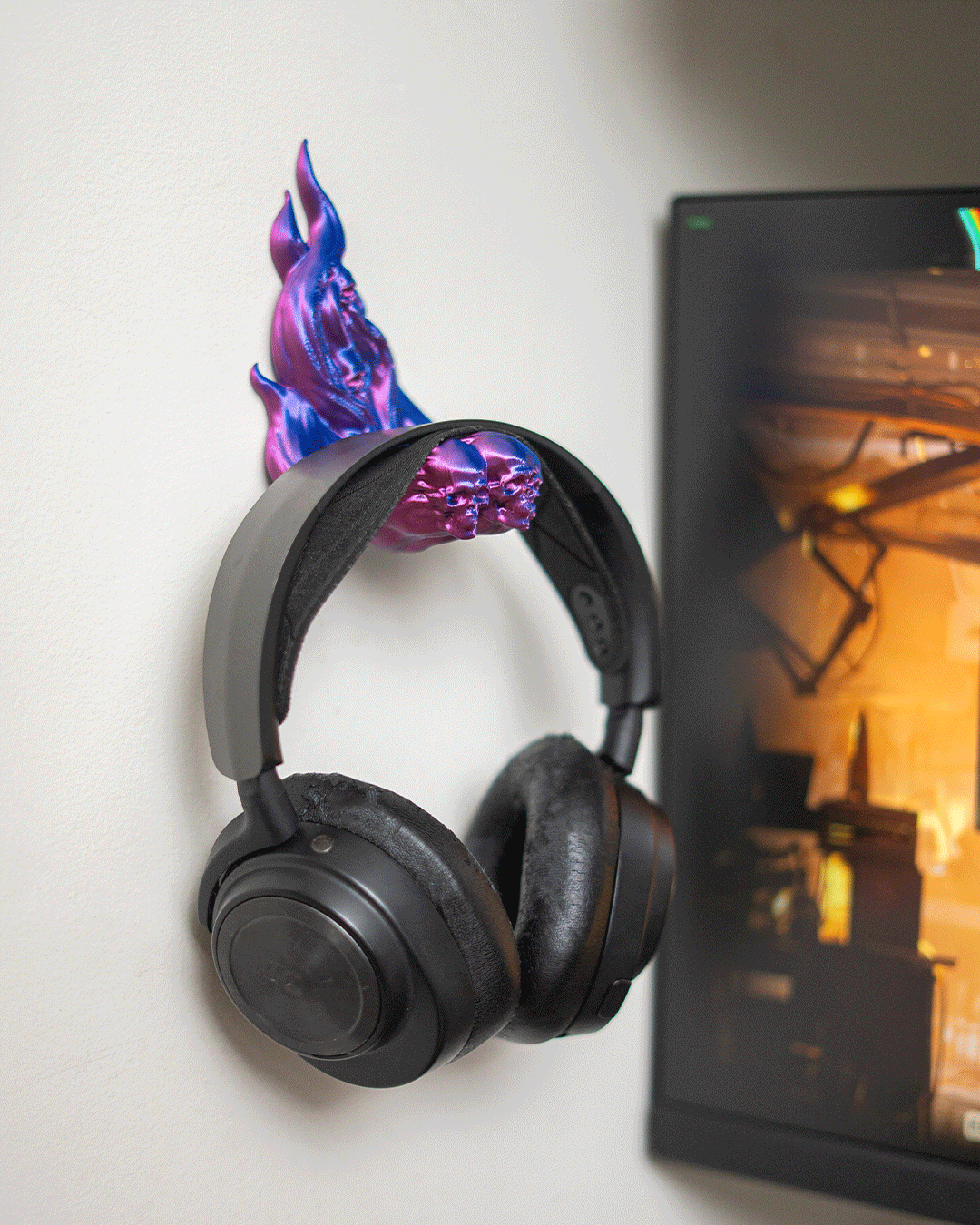 Skullflame Wand-Headset-Halter in Pink-Lila mit aufgehängtem Over-Ear-Kopfhörer, an heller Wand neben einem Monitor mit Spielszene im Hintergrund.
