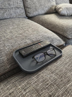 Dunkelgraues geriffeltes Sofatablett mit Fernbedienung und schwarzer Brille, auf grauer Couch-Armlehne, kein Kaffeegeschirr im Bild