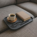 Dunkelgraues Sofatablett mit Kaffeetasse auf Holzuntersetzer, beigefarbenem Buch, schwarzer Brille und Kugelschreiber, auf grauer Couch-Armlehne