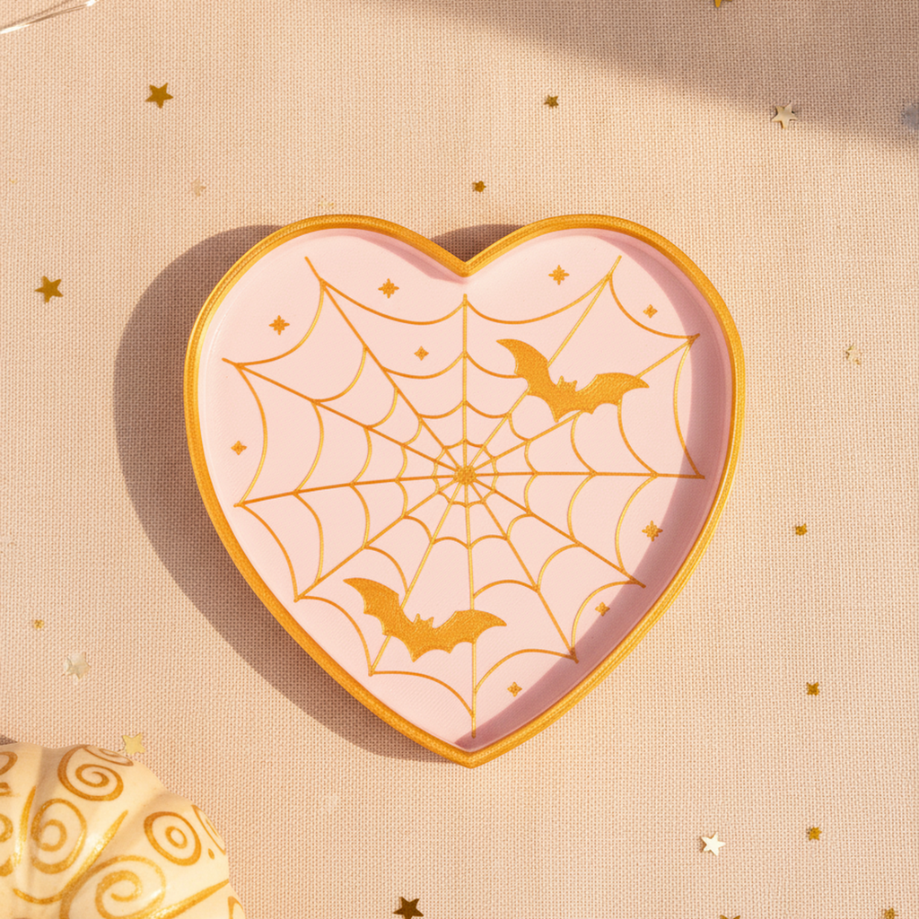 Herzförmige Spinnennetz-Deko-Schale in Rosa und Gold mit zwei Fledermäusen, Draufsicht auf blassrosa Textiluntergrund mit goldenen Sternkonfetti und Lichterkette