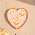 Herzförmige Spinnennetz-Deko-Schale in Rosa und Gold mit zwei Fledermäusen, Draufsicht auf blassrosa Textiluntergrund mit goldenen Sternkonfetti und Lichterkette
