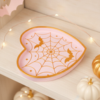 Spinnennetz-Herz-Schale in Rosa-Gold auf einem cremefarbenen Holzregal zwischen Mini-Kürbissen, Lichterkette und pastellfarbenen Papier-Fledermäusen