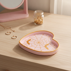 Spinnennetz-Herz-Schale in Rosa-Gold auf einer hellen Kommode neben goldenen Ohrringen, einem Ring und einer Halskette, im Hintergrund ein rosa Spiegel