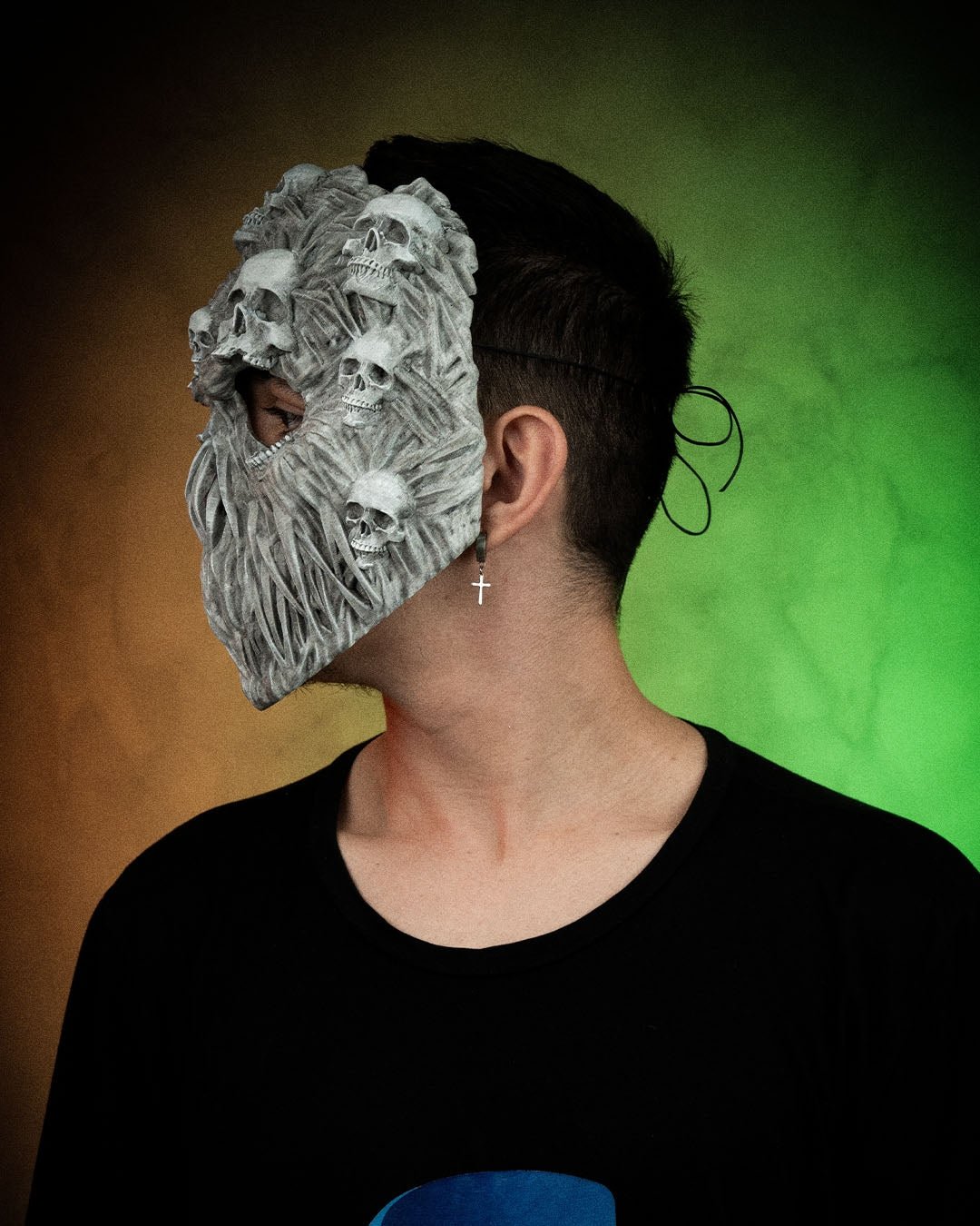 Ghoul-Totenkopf-Maske in Grau getragen von einer Person, Seitenprofil mit Ohrring sichtbar, bunter Farbverlauf-Hintergrund