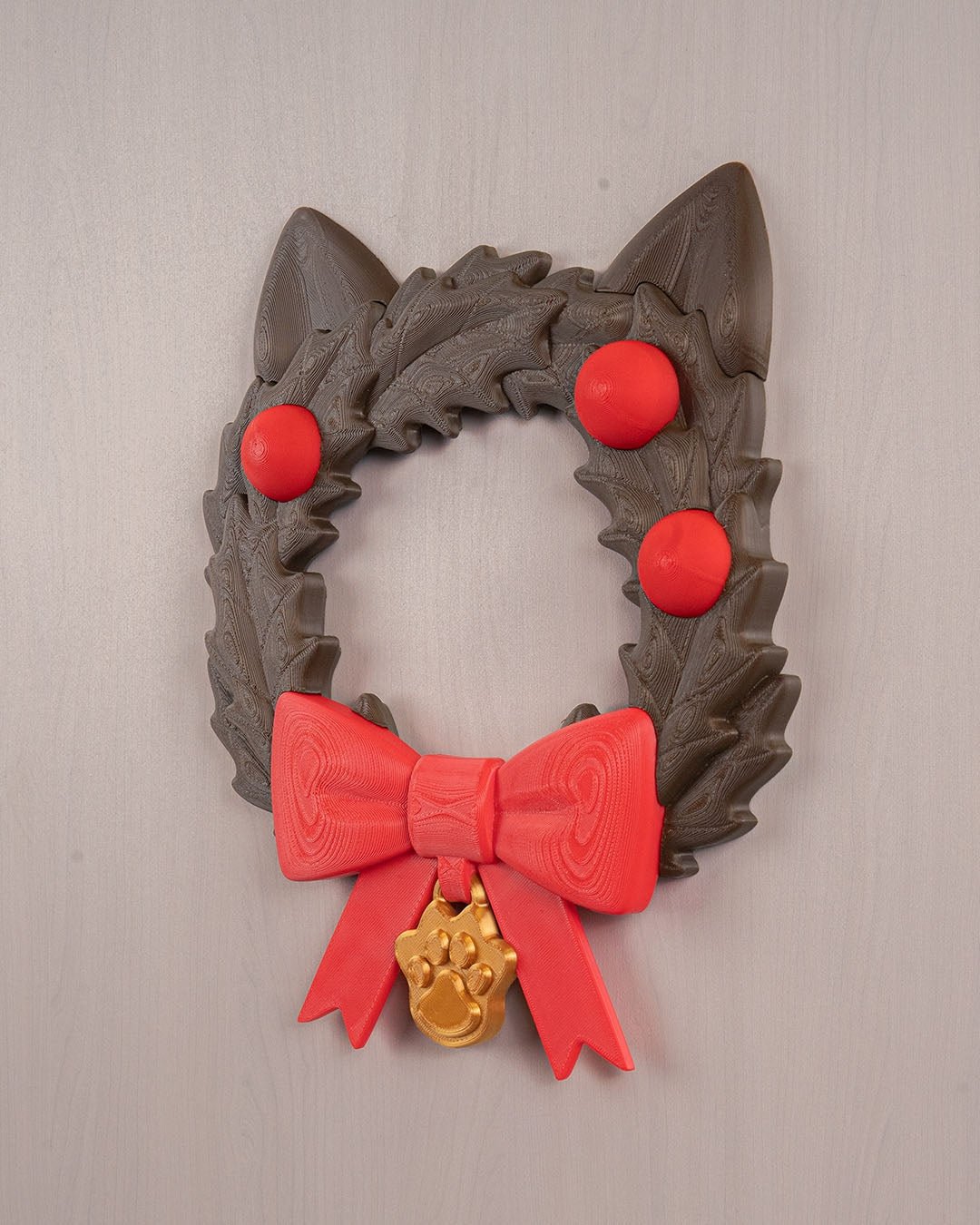 Weihnachtskranz in Katzenkopf-Form mit Katzenohren, roten Kugeln, roter Schleife und goldenem Pfotenanhänger, Frontalansicht an heller grauer Wand mit sichtbarer Holzmaserung