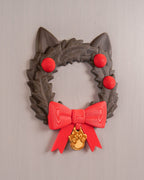 Weihnachtskranz in Katzenkopf-Form mit Katzenohren, roten Kugeln, roter Schleife und goldenem Pfotenanhänger, Frontalansicht enger ausgeschnitten an heller grauer Wand