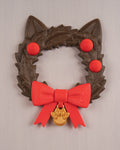 Weihnachtskranz in Katzenkopf-Form mit Katzenohren, roten Kugeln, roter Schleife und goldenem Pfotenanhänger, Frontalansicht an heller grauer Wand