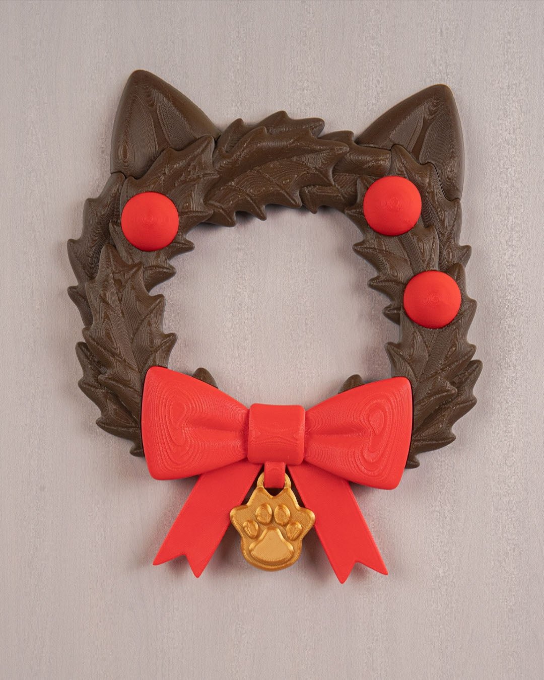 Weihnachtskranz in Katzenkopf-Form mit Katzenohren, roten Kugeln, roter Schleife und goldenem Pfotenanhänger, Frontalansicht an heller grauer Wand