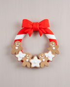 Weihnachtstürkranz mit Lebkuchenmännern, weißen Zuckerguss-Sternen und roter Zuckerstangen-Schleife, Frontalansicht an heller beige-rosa Wand