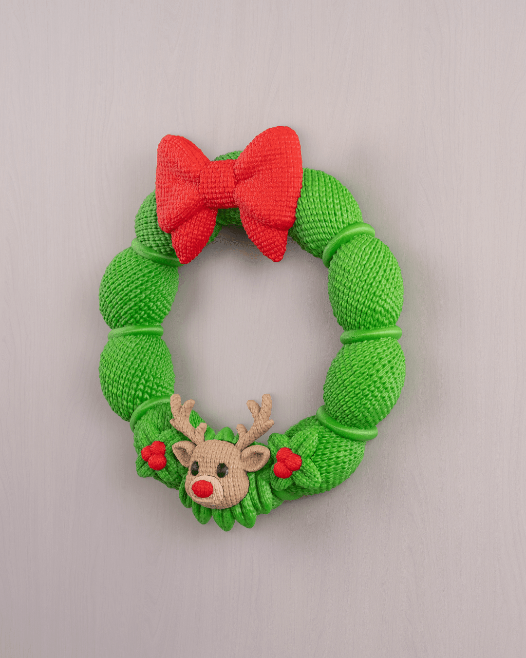 Weihnachtskranz aus grüner Strickoptik mit rotem Schleifen-Band und Rentier-Applikation mit roter Nase, Frontalansicht an heller grauer Wand