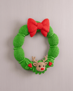 Weihnachtskranz aus grüner Strickoptik mit rotem Schleifen-Band und Rentier-Applikation, Frontalansicht an heller grauer Wand, leicht anderer Bildausschnitt
