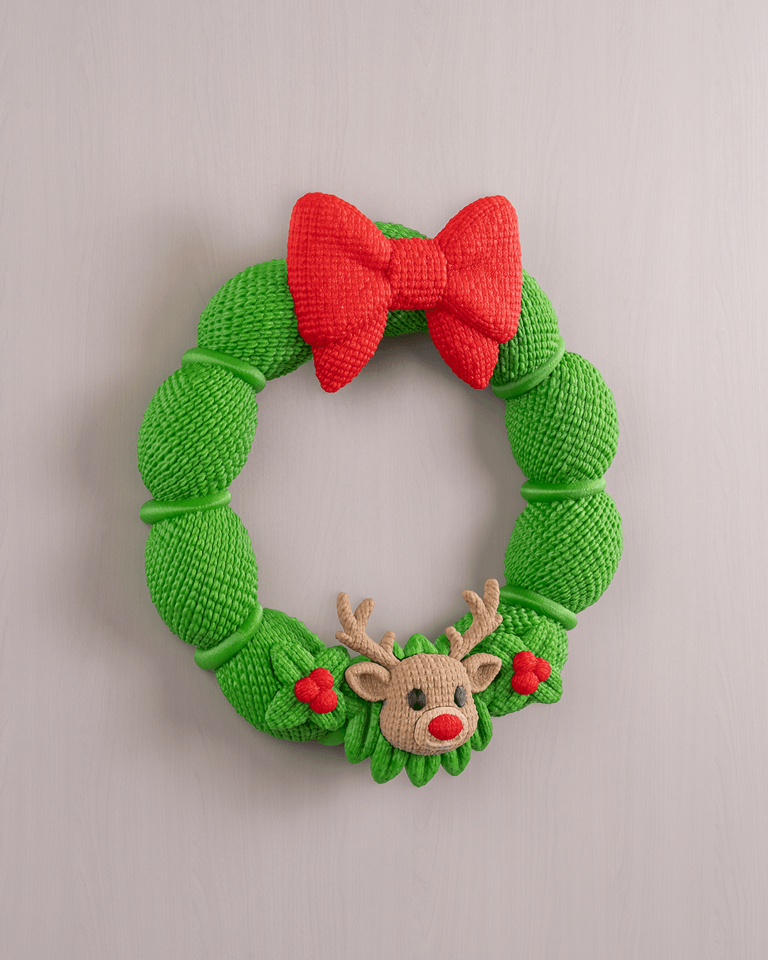 Weihnachtskranz aus grüner Strickoptik mit rotem Schleifen-Band und Rentier-Applikation, Frontalansicht an heller grauer Wand, leicht anderer Bildausschnitt