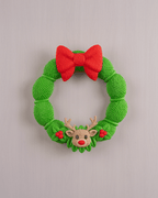 Weihnachtskranz aus grüner Strickoptik mit rotem Schleifen-Band und Rentier-Applikation mit roter Nase, Frontalansicht an heller grauer Wand