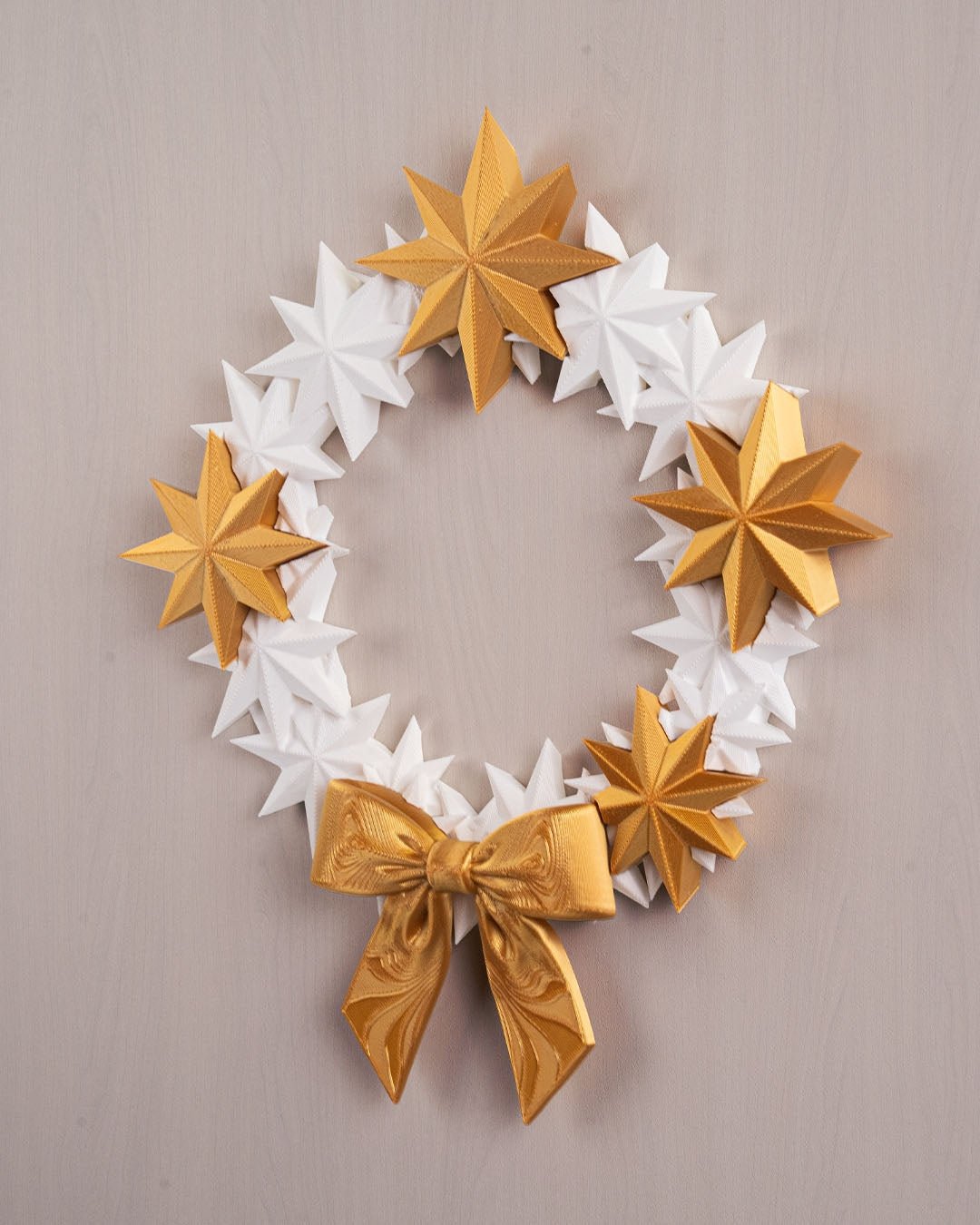 Sternenkranz aus weißen und goldenen 3D-gedruckten Sternen mit goldener Schleife, Frontalansicht an heller Wand