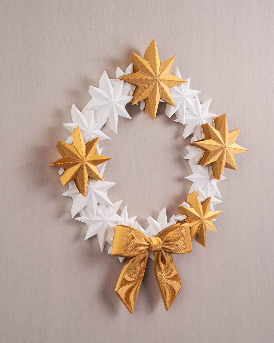 Sternenkranz aus weißen und goldenen 3D-gedruckten Sternen mit goldener Schleife, Frontalansicht an heller Wand, leicht anderer Bildausschnitt
