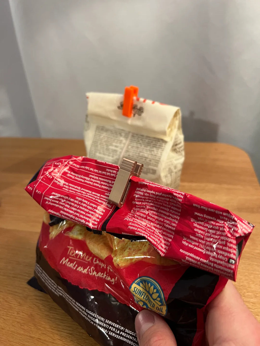 Kupferfarbener Tütenclip an einer aufgerollten Chipstüte, im Hintergrund weitere Tüte mit orangem Clip
