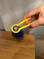 Gelber Flaschenöffner umschließt den Deckel einer blauen Trinkflasche, seitliche Ansicht in der Hand