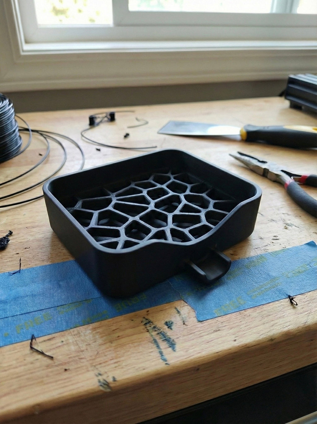 Unlackierter schwarzer 3D-Druck-Prototyp der Voronoi-Seifenschale mit sichtbarem Zellstruktur-Gitter, leer ohne Seife, auf einer Werkbank mit Drahtspulen, Zange und Klebeband vor einem Fenster.