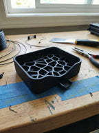 Unlackierter schwarzer 3D-Druck-Prototyp der Voronoi-Seifenschale mit sichtbarem Zellstruktur-Gitter, leer ohne Seife, auf einer Werkbank mit Drahtspulen, Zange und Klebeband vor einem Fenster.
