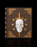 Würfelbox in Buchform geschlossen, braunes Lederdesign mit goldenen Ecken-Ornamenten, weißem Totenschädel-Relief und goldenem Dolch, Frontalansicht vor schwarzem Hintergrund