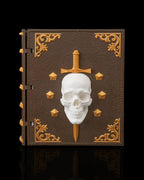 Würfelbox in Buchform geschlossen, braunes Lederdesign mit goldenen Ecken-Ornamenten, weißem Totenschädel-Relief und goldenem Dolch, Frontalansicht vor schwarzem Hintergrund