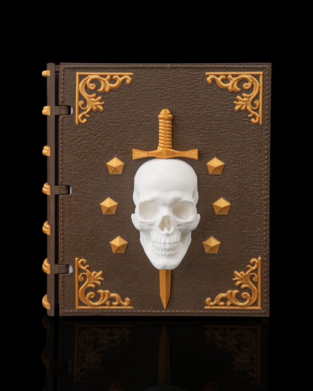 Würfelbox in Buchform geschlossen, braunes Lederdesign mit goldenen Ecken-Ornamenten, weißem Totenschädel-Relief und goldenem Dolch, Frontalansicht vor schwarzem Hintergrund