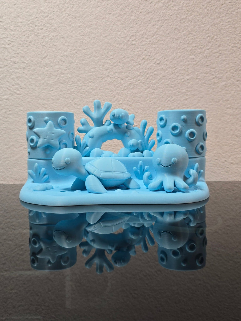 Eisblauer Zahnbürstenhalter mit Schildkröte, Oktopus, Seestern und Korallenbogen in direkter Frontalansicht, leer ohne Zahnbürste, auf dunkler Ablage vor strukturierter Wand
