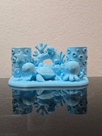 Eisblauer Zahnbürstenhalter mit Schildkröte, Oktopus, Seestern und Korallenbogen in direkter Frontalansicht, leer ohne Zahnbürste, auf dunkler Ablage vor strukturierter Wand