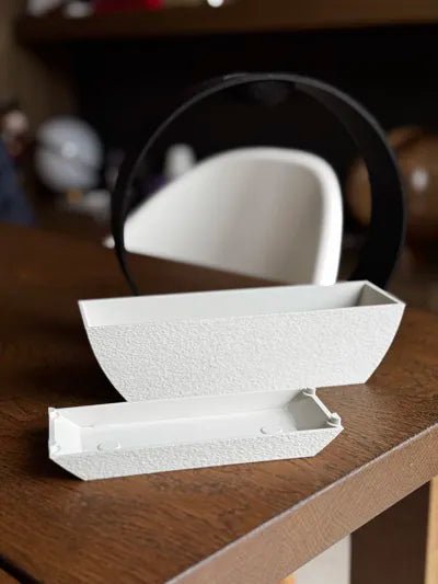 Zerlegte ZenRain Vase: weiße, steinstrukturierte Pflanzschale und ein kleinerer schmaler weißer Wassereinsatz liegen einzeln nebeneinander auf einem Holztisch, unbepflanzt, schwarzer Ring-Rahmen unscharf im Hintergrund.