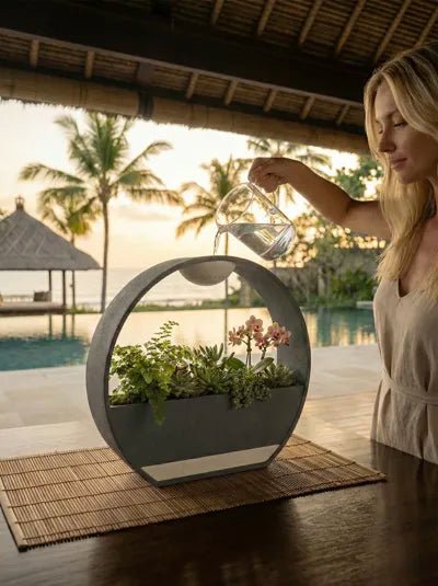 ZenRain Vase in Lifestyle-Szene: Frau gießt Wasser aus einer Glaskaraffe in die runde Vase mit Basilikum, Rosmarin, Minze und rosafarbener Orchidee, auf Bambusmatte auf einem Holztisch, im Hintergrund Pool und Palmen bei Sonnenuntergang.